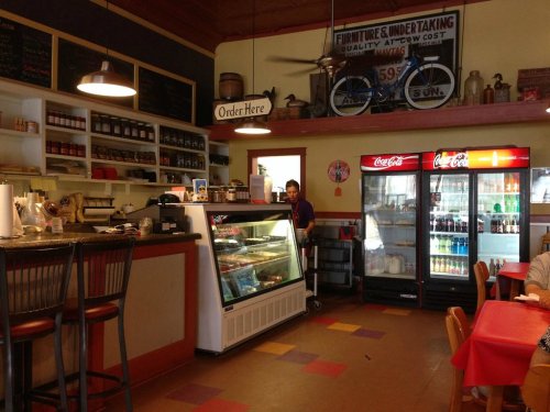 Cozy café interior with a counter, display case, and drink refrigerators; a staff member is at the register.