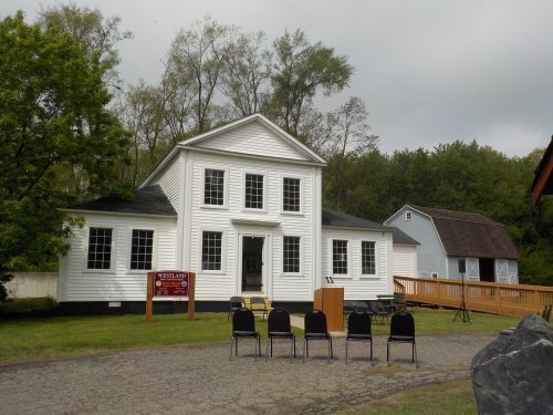 A white historic building with large windows, surrounded by trees, and empty chairs set up for an event outside.