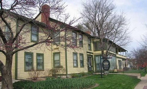 A two-story yellow house with green trim, surrounded by trees and flowers, featuring a sign in front.