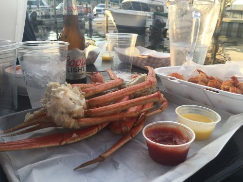 A seafood platter featuring crab legs, dipping sauces, and drinks by the water, with boats in the background.
