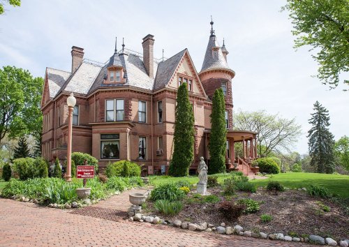A large, historic red brick mansion with a turret, surrounded by greenery and decorative statues in the garden.