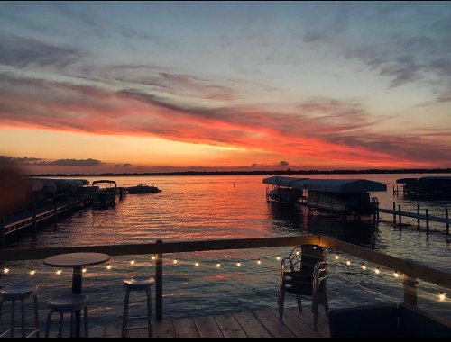 A serene sunset over a calm waterway, with boats docked and string lights illuminating a wooden deck.