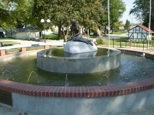 A circular fountain with a statue on a rock, surrounded by water and greenery in a park setting.