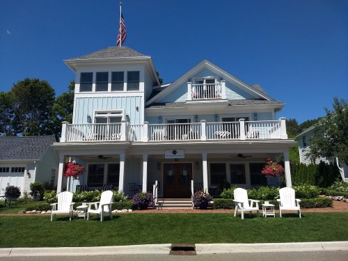 A large blue house with a flag, featuring balconies, white chairs, and colorful flowers in a sunny setting.