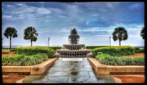 A decorative fountain shaped like a pineapple, surrounded by lush greenery and palm trees, under a cloudy sky.