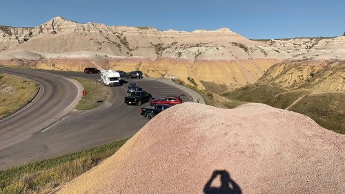 A winding road curves through colorful rock formations, with parked vehicles along the roadside and a shadow in the foreground.