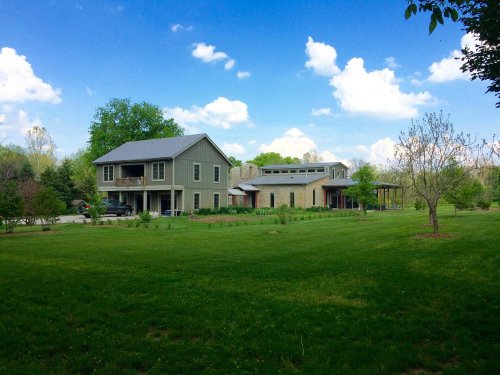 A spacious green lawn with two modern houses under a blue sky with fluffy clouds. Trees and shrubs surround the area.