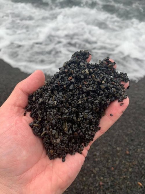 A hand holding a handful of dark, shiny pebbles or gravel against a backdrop of ocean waves.