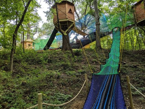 A colorful rope bridge leads to a treehouse in a wooded area, surrounded by greenery and a netted play structure.
