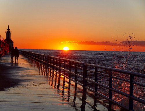 Silhouetted figures walk along a pier at sunset, with waves splashing and vibrant colors in the sky.