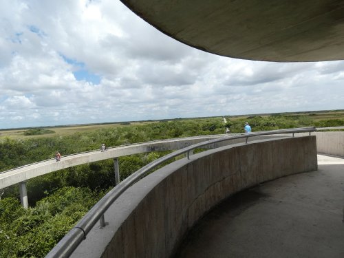 View from a curved concrete observation deck overlooking lush greenery and a cloudy sky. People walk along the path.