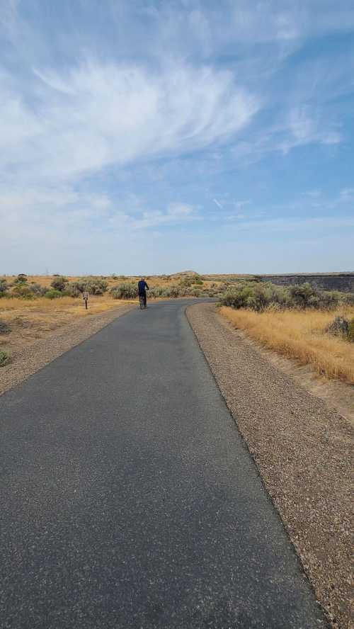 A person walking on a winding path through a dry, grassy landscape under a blue sky with scattered clouds.