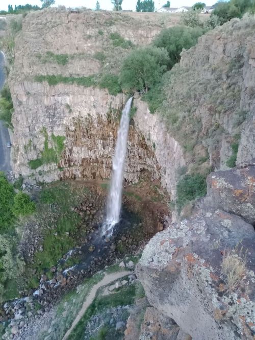 A waterfall cascading down rocky cliffs surrounded by greenery and a winding path below.