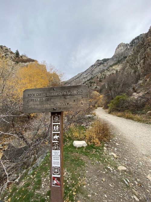 Trail sign at Rock Canyon with directions, surrounded by autumn foliage and mountains in the background.