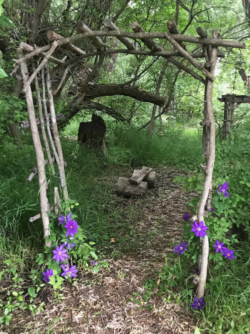 A rustic wooden arch adorned with purple flowers, leading into a lush, green forest path.