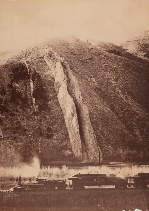 A vintage photograph of a steam train near a mountain, with a river and rocky terrain in the background.