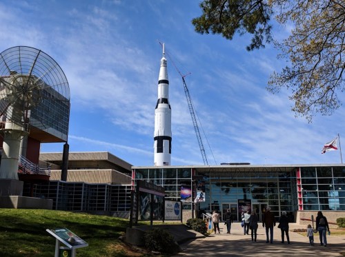 A rocket stands tall outside a modern building, with visitors walking towards the entrance under a blue sky.