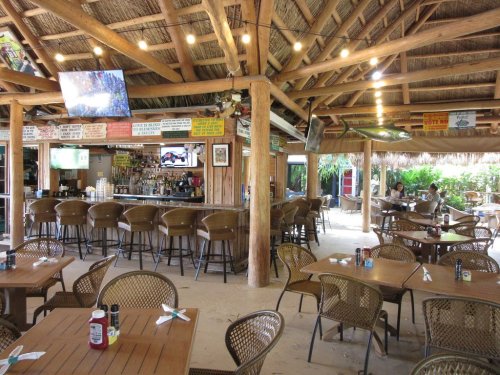 A rustic bar and dining area with wooden beams, tables, and a thatched roof, featuring TVs and tropical decor.