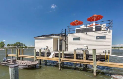 A modern houseboat with red umbrellas on the deck, docked by a wooden pier on a calm waterway.