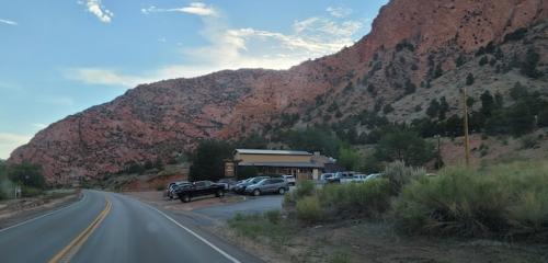 A scenic view of a road alongside a rocky mountain, with a building and parked cars visible.