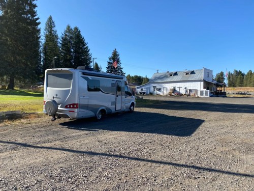 A white RV parked on gravel near a rustic building surrounded by trees and clear blue skies.