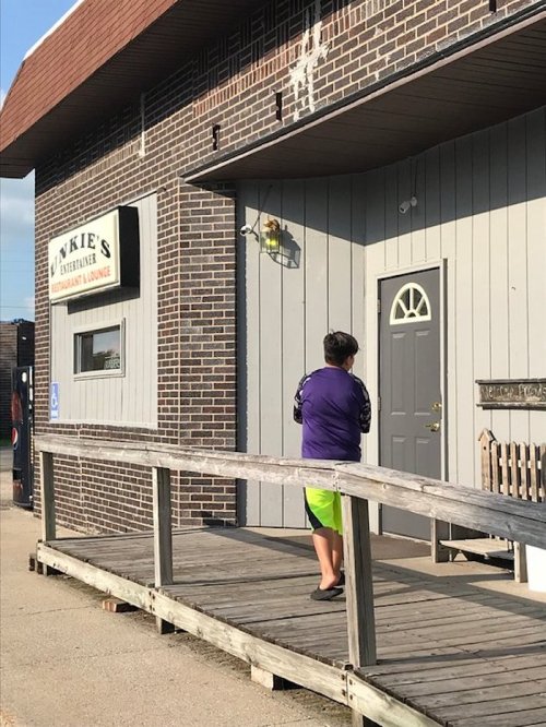 A person in a purple shirt and green shorts stands on a wooden ramp in front of a restaurant lounge with a sign.