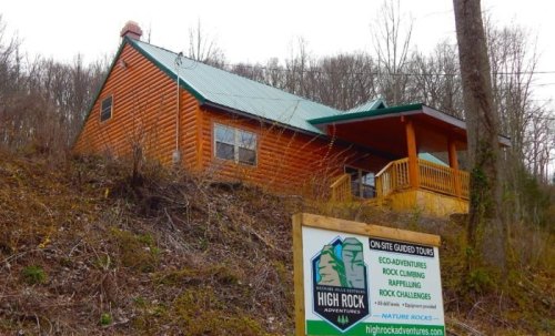 A wooden cabin with a green metal roof, surrounded by trees, next to a sign for High Rock Adventures.