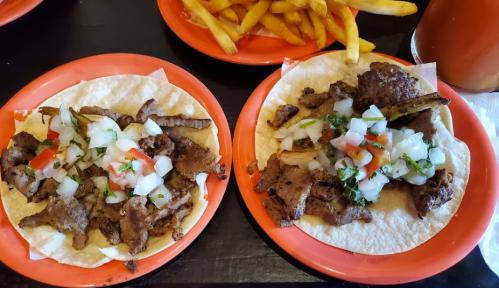 Two plates of tacos filled with seasoned meat and topped with diced onions, tomatoes, and cilantro, served with fries.