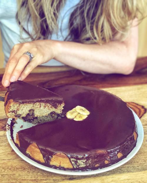 A person slices a chocolate-covered cake with banana slices on top, sitting at a wooden table.