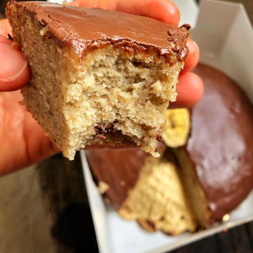 A hand holding a piece of chocolate-covered cake, with a larger cake in the background, partially sliced.