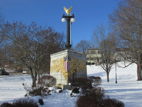 A snow-covered monument featuring an eagle and an ornate shield, surrounded by bare trees and a clear blue sky.