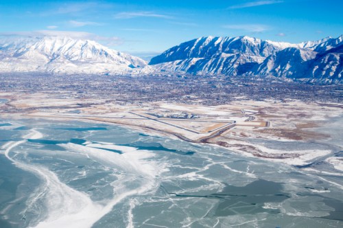 Aerial view of snow-covered mountains and a frozen lake, with a city in the valley below under a clear blue sky.