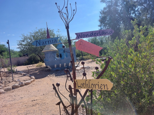 A whimsical signpost with directions to Narnia, Wonderland, and Lothlorien, near a colorful, castle-like structure.