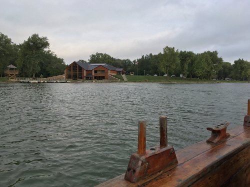 A calm lake scene with a wooden building and trees in the background, viewed from a boat.