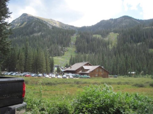 A scenic view of a mountain lodge surrounded by trees and parked cars, with mountains in the background.