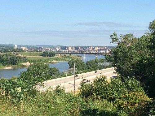 A scenic view of a river with a bridge, city skyline in the background, and lush greenery in the foreground.