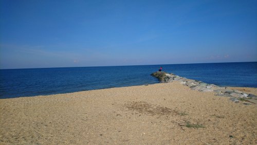 A person stands on a rocky jetty extending into a calm blue sea under a clear sky. Sandy beach in the foreground.