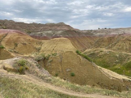 A scenic view of colorful, layered hills under a cloudy sky, with grassy areas and a winding path in the foreground.