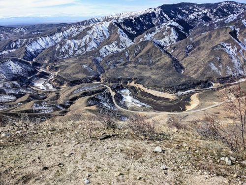 A panoramic view of rugged, snow-capped mountains and winding river valleys under a clear blue sky.