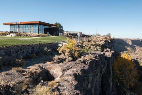 A modern building sits atop a rocky cliff, surrounded by greenery and autumn-colored trees against a clear blue sky.