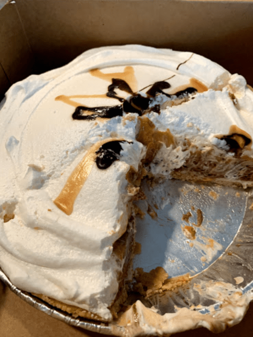 A partially sliced dessert with whipped cream topping, chocolate drizzle, and a graham cracker crust in a cardboard box.