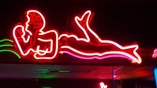 Neon sign of a mermaid lounging, glowing in red and pink against a dark background.