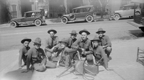 A group of nine men in military uniforms and hats, posing with equipment, with vintage cars in the background.