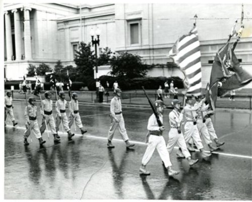A group of soldiers marching in a parade, carrying flags, with a historic building in the background.