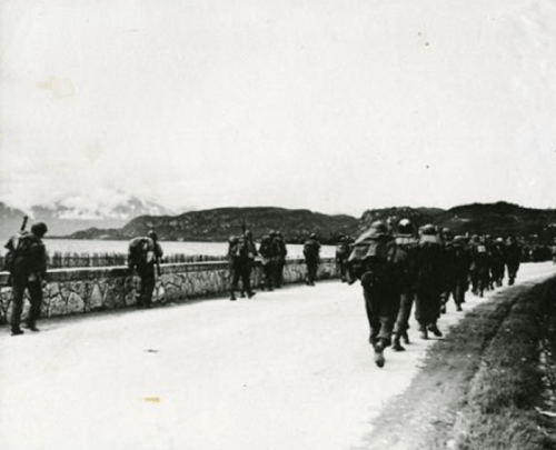 Black and white photo of soldiers marching along a coastal road with mountains in the background.