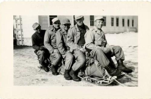 Five soldiers sitting on a sled, smiling, with snow in the background and buildings behind them.
