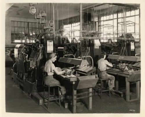 Two workers operate large machinery in a factory setting, with rows of equipment and overhead lighting.