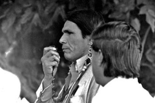 A man with long hair and earrings speaks into a microphone, while another person is partially visible beside him.
