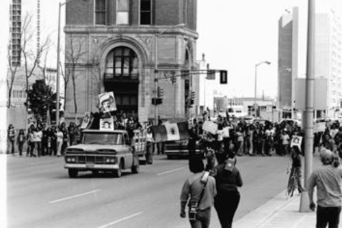 A black and white photo of a protest march with a crowd, flags, and a truck in an urban setting.