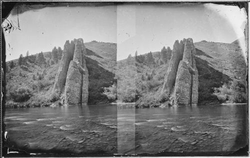A black-and-white photo of tall rock formations beside a river, surrounded by trees and hills in the background.
